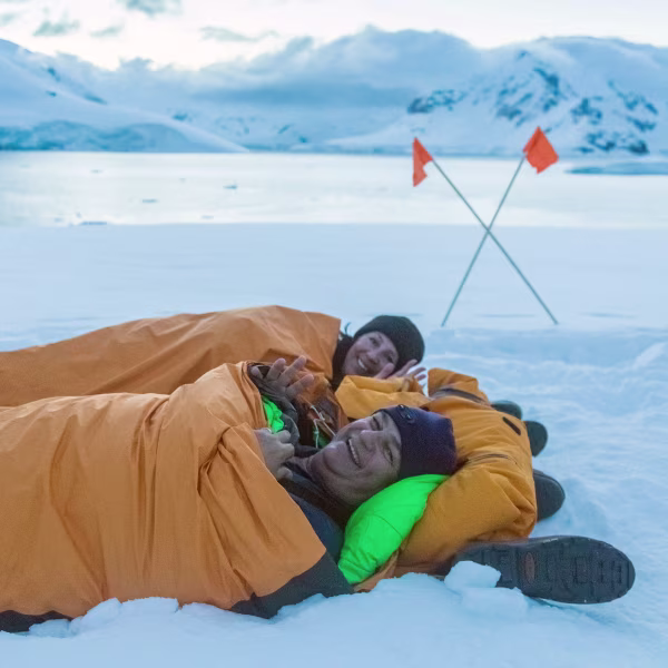 Dois campistas em sacos de bivaque laranja sobre a neve, ao lado de bandeiras de sinalização em Leith Cove, Antártica