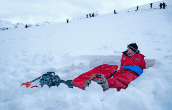 Uma pessoa acampando em um saco de dormir vermelho dentro de uma trincheira escavada na neve, com outras caminhando pela crista acima